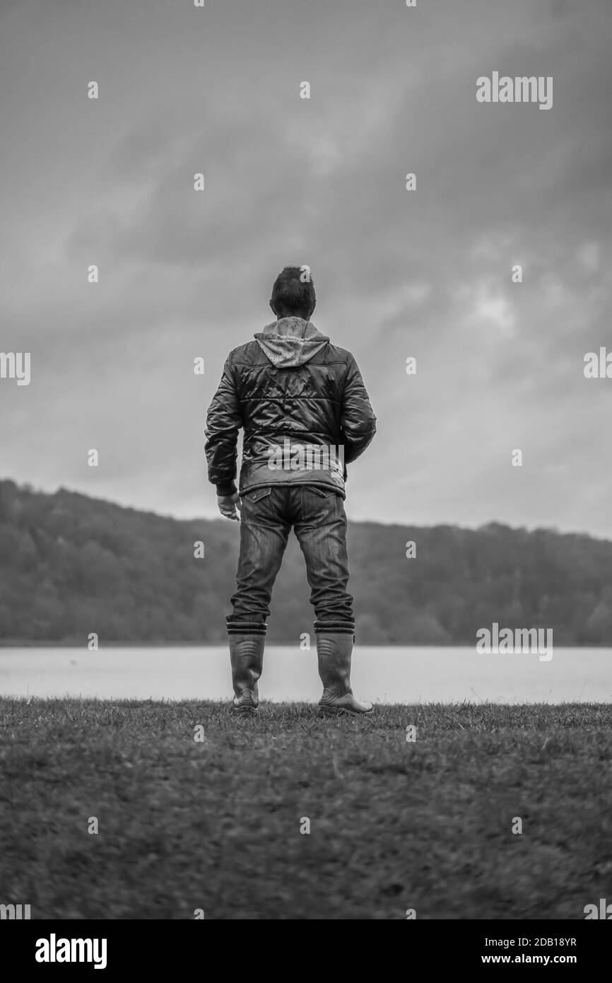 Black and white, rear view of isolated man in coat standing by a lake ...