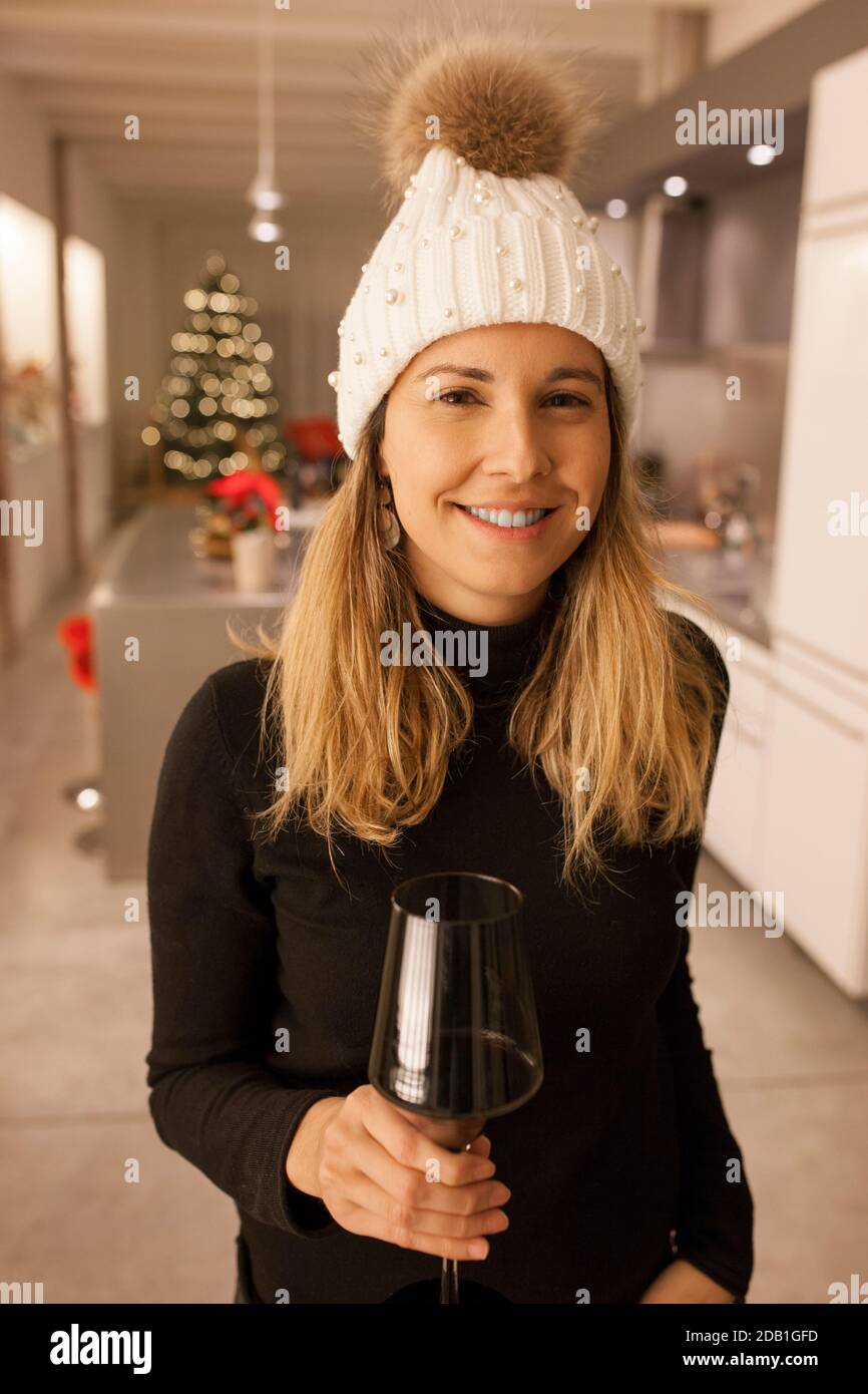 Beautiful smiling woman wearing white beani and holding up a cup of red wine in the kitchen with christmas tree background Stock Photo