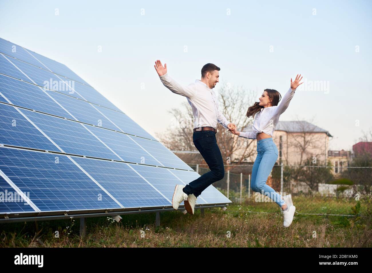 Young family jumping holding hands near the house with solar panels. Guy and girl are dressed in jeans, shirts and white sneakers are looking at each other. Solar energy concept image Stock Photo