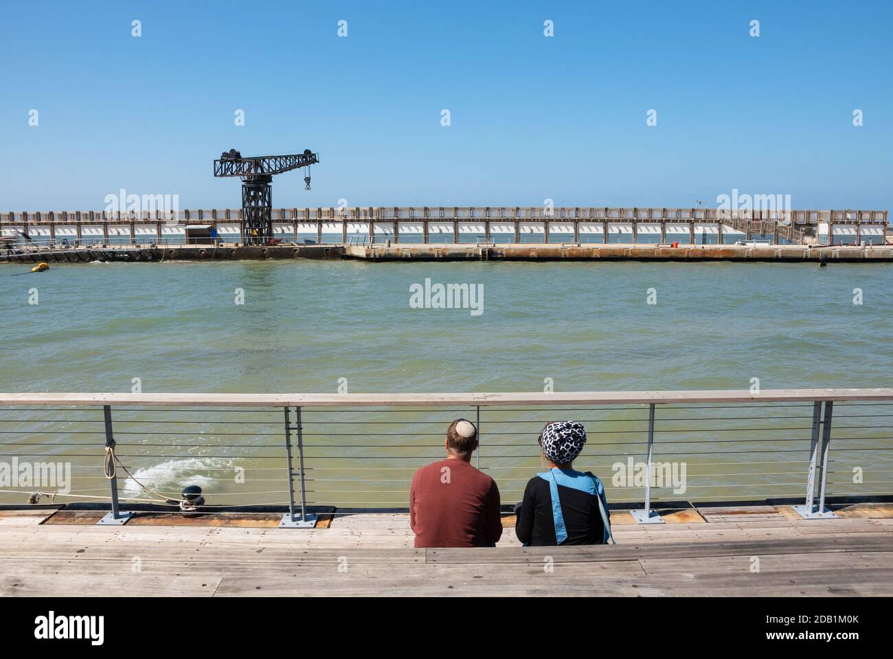 Jewish young couple (man wearing kippah and woman with head scarf) relaxing in sunny spring day at Tel Aviv port, Israel. Back view; unrecognizable pe Stock Photo
