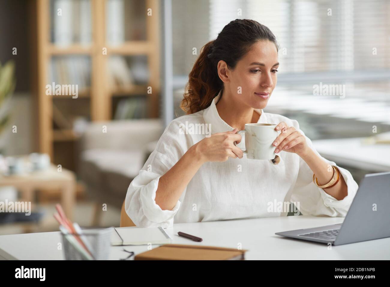 Portrait of elegant young woman looking at laptop screen and drinking coffee while enjoying work from home in minimal white interior, copy space Stock Photo