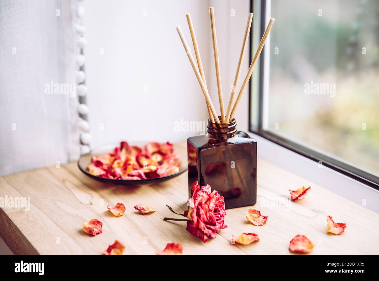 Brown glass bottle container with wood stick diffusers on home wooden window sill with beautiful dry pink rose petals for decoration. Stock Photo