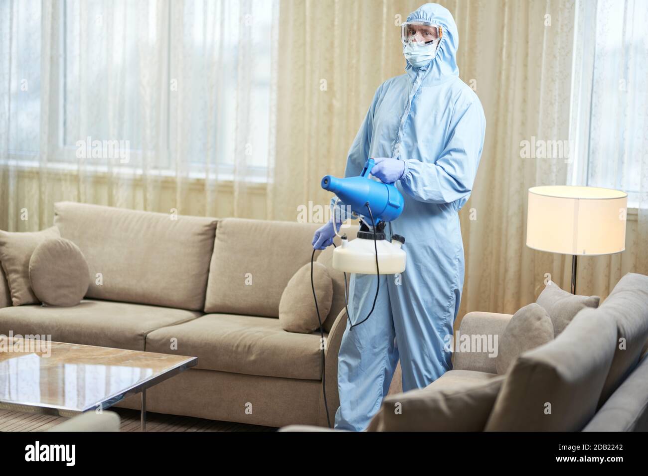 Worker wearing protective suit and glasses standing in hotel room and using disinfectant. Coronavirus and quarantine concept Stock Photo