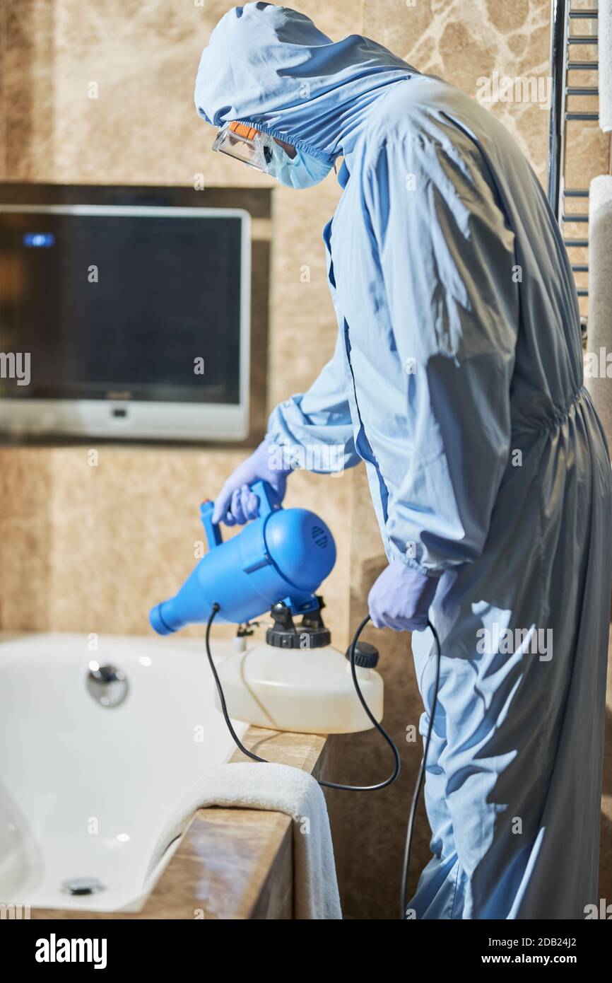 Side view of of worker in biohazard suit using disinfectant in hotel bathroom. Coronavirus and quarantine concept Stock Photo