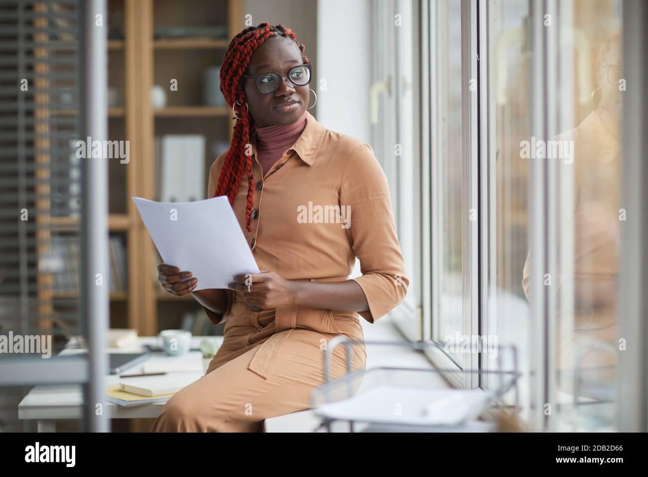 Portrait of contemporary African-American woman looking away pensively and holding document while leaning on window in office, copy space Stock Photo