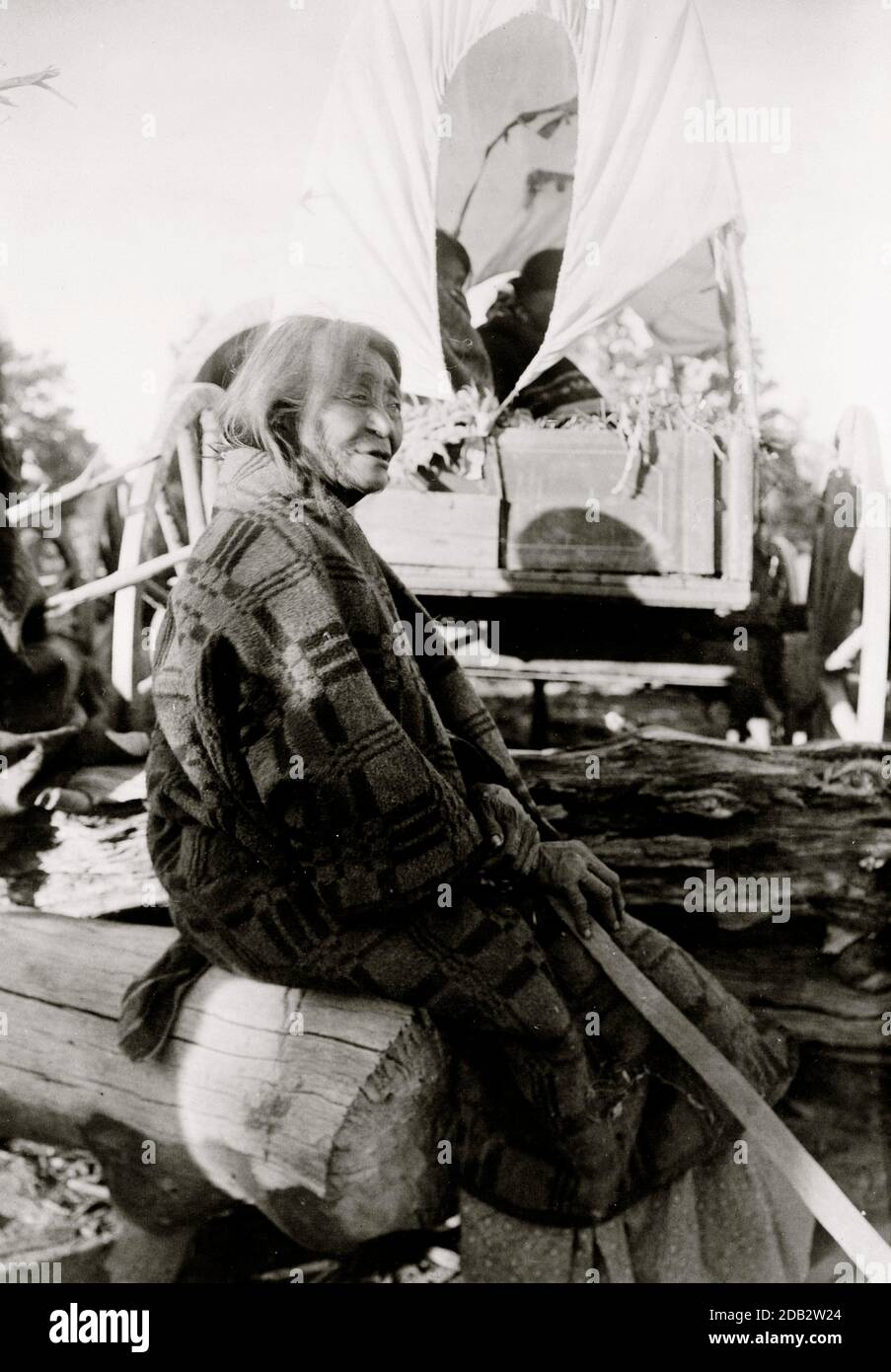 Old Navajo Indian woman, seated on log, back of covered wagon in background.. Stock Photo