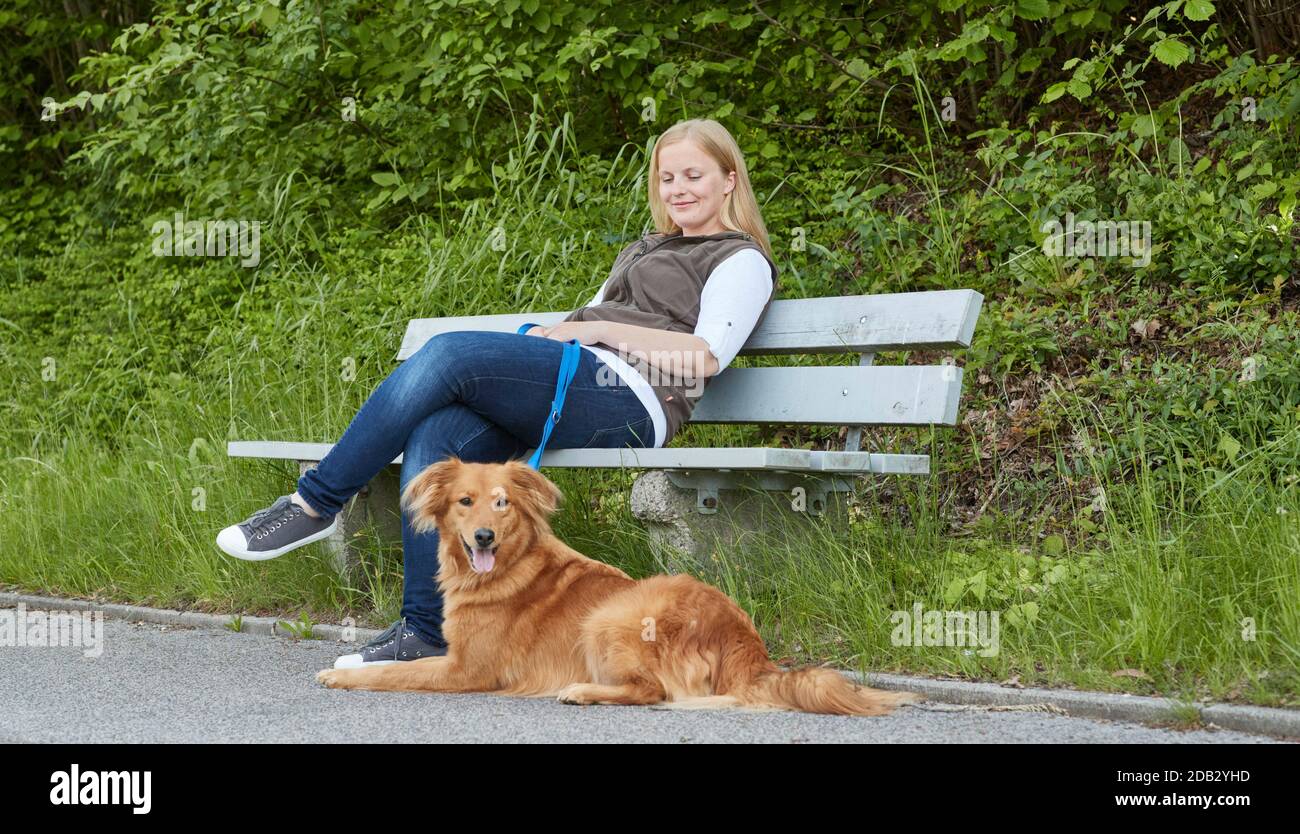 Woman sits on park bench, mixed breed dog lying in front of it. Not for animal guide books until 9/2022 Stock Photo