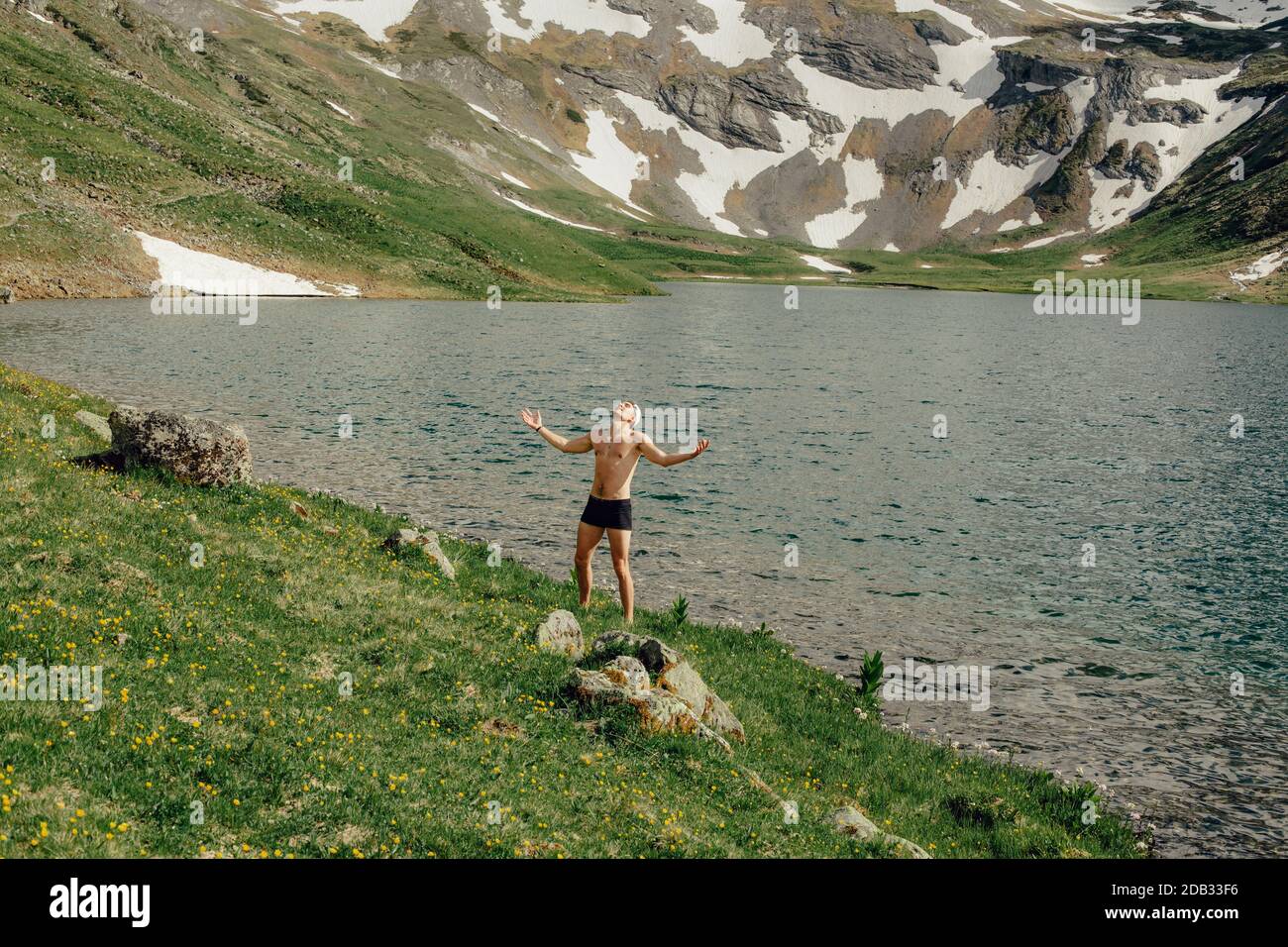 Happy young male tourist in a bathing suit is happy that he has reached his goal and reached the mountain lake enjoying the beautiful grand nature on Stock Photo