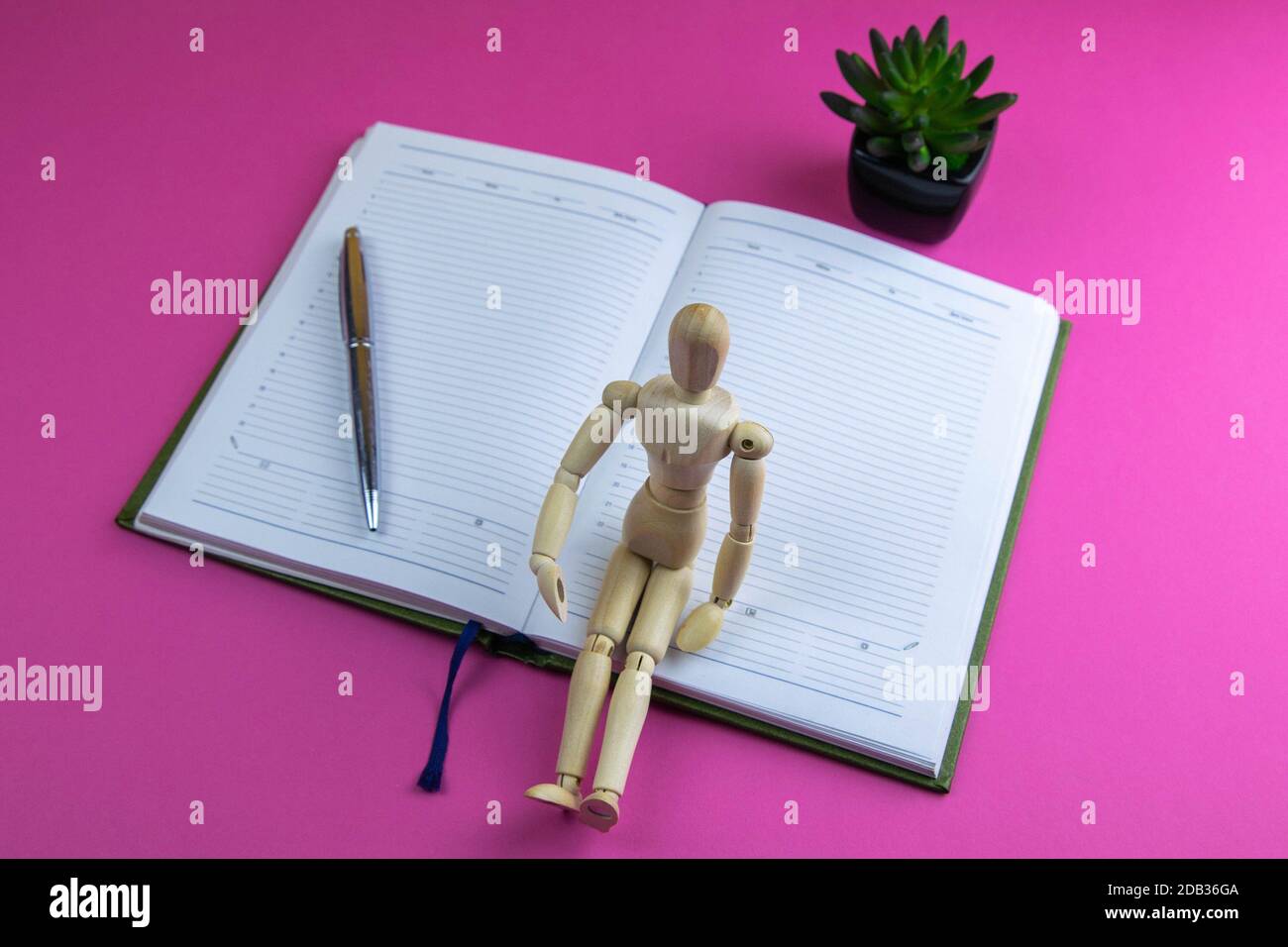 The figurine of a little wooden man sits on an open notebook, next to it stands a silver pen and an artificial green flower. Everything stands on a pi Stock Photo