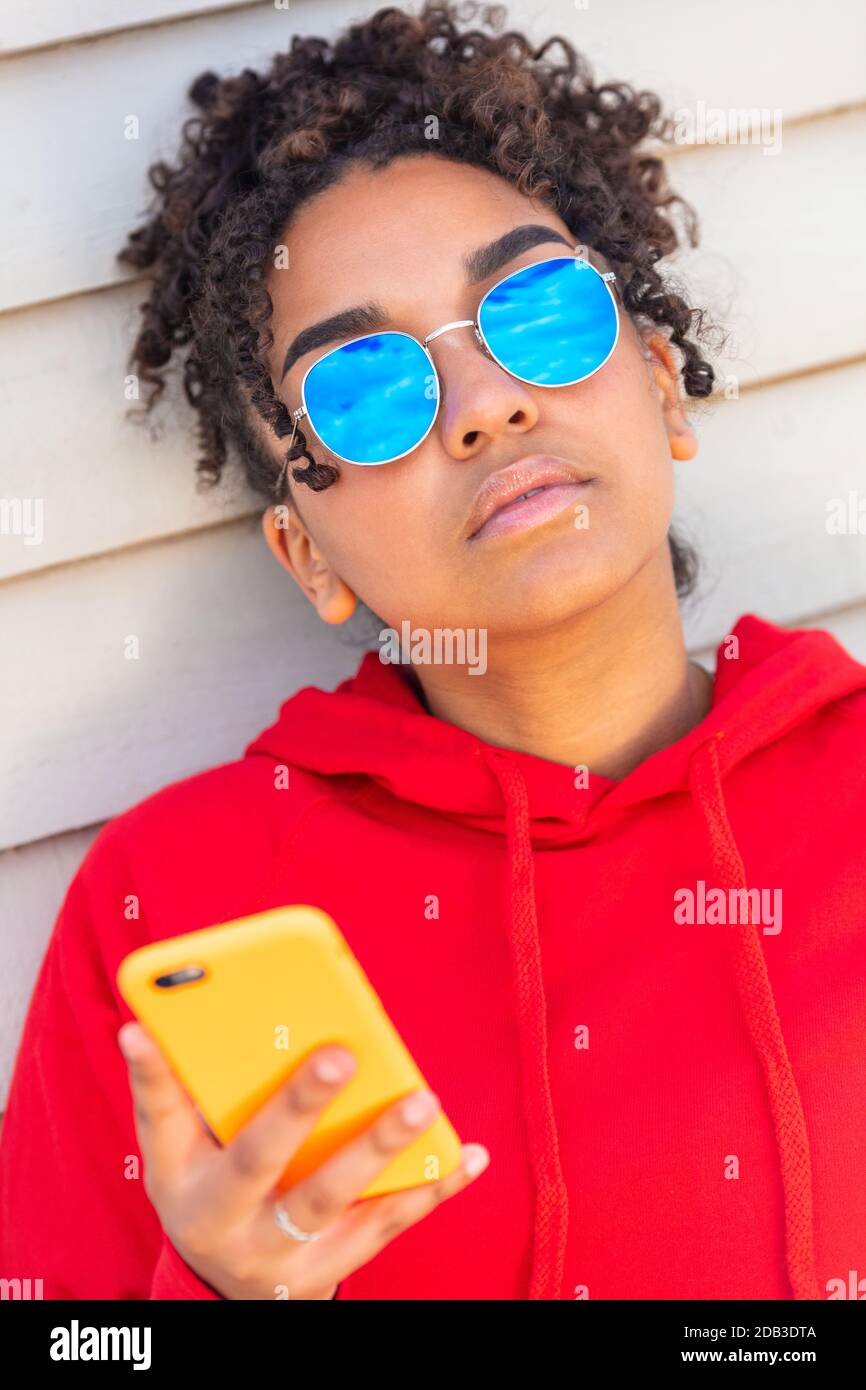 Girl teenager cool teen mixed race biracial African American female young woman wearing blue sunglasses and a red hoodie using cell phone on vacation Stock Photo