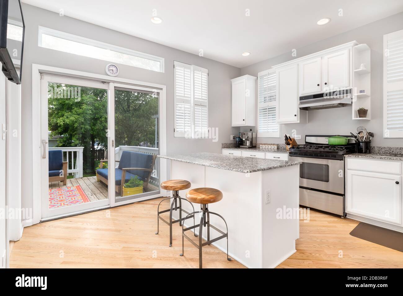 A white kitchen with grey marble countertops, stainless steel appliances, and wooden seat bar stools sitting at the island. Stock Photo