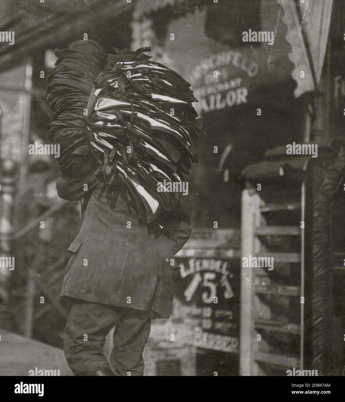 Man loaded down with heavy bundle of vests that he had been carrying for many blocks, stopping to rest, occasionally, when he would drop the bundle down onto whatever boxes or railings were available, even though they were dirty. Stock Photo