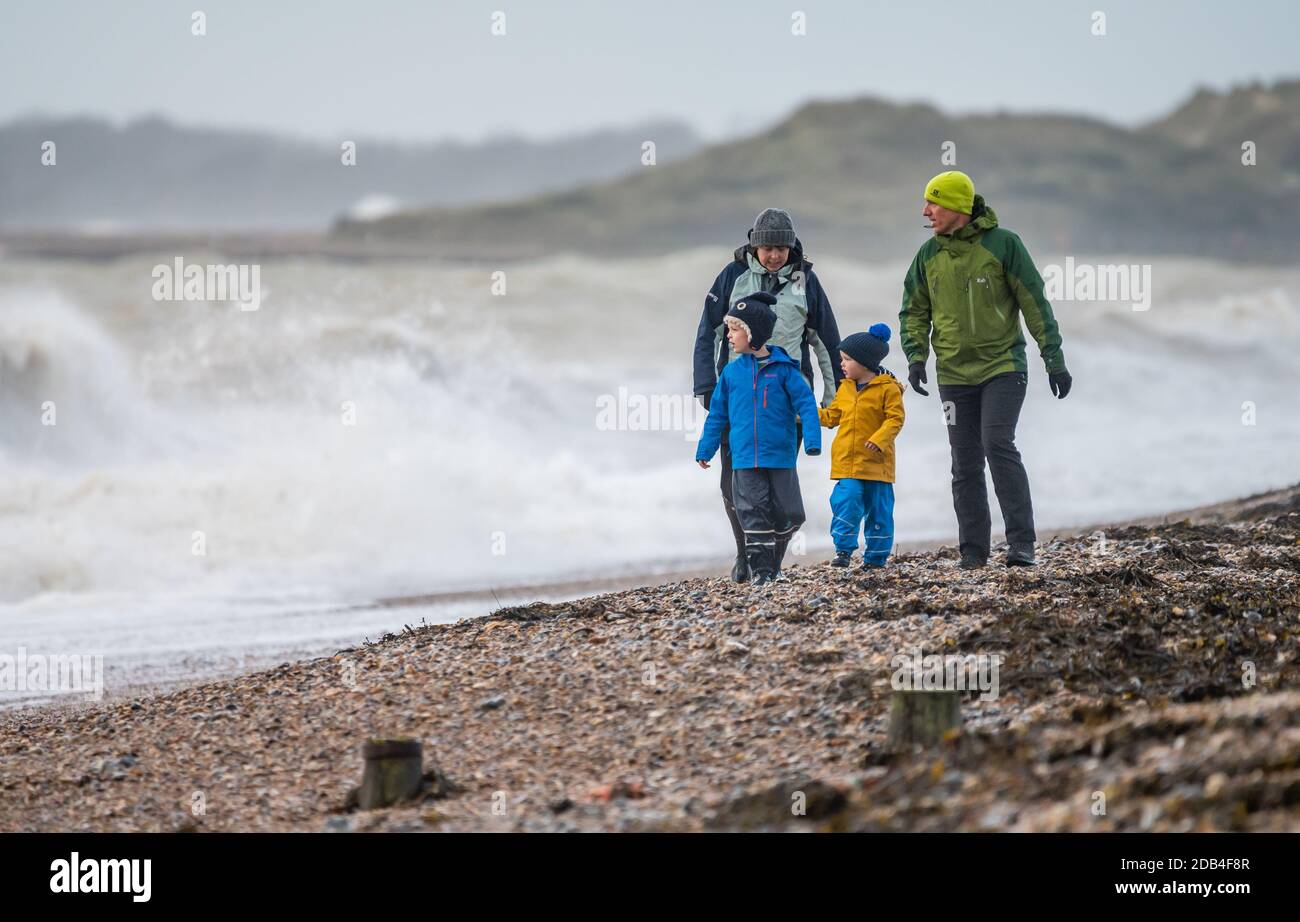 Family of 2 adults and 2 children walking along a beach with rough sea, strong wind, generally poor weather, in the UK. Coastal walk. Stock Photo