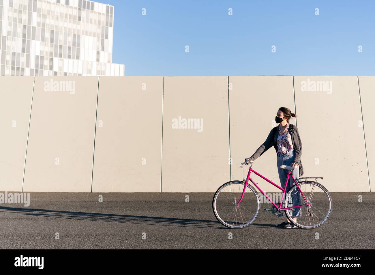 young woman with protective mask standing with a pink retro bike at city, concept of active lifestyle, protection against covid and sustainable urban Stock Photo