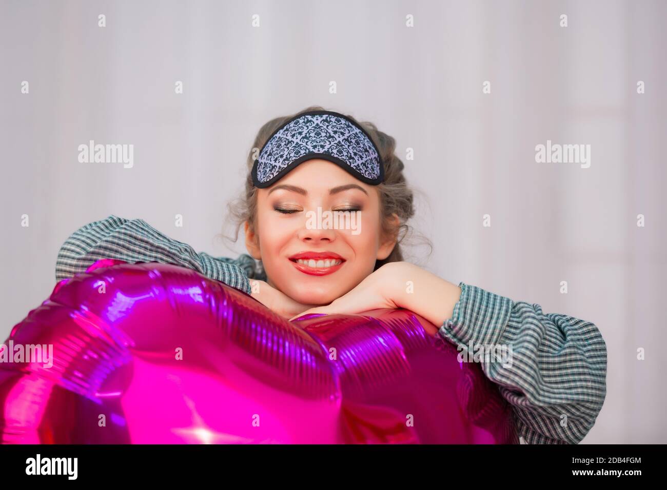 Young woman in sleep mask hugs pink heart balloon and smiles with closed eyes Stock Photo