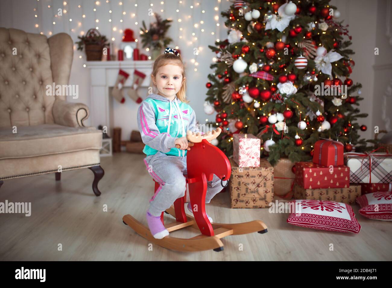 A little girl in a tracksuit sits on a wooden red moose rocking chair near a Christmas tree. Christmas, new year, holiday decorations Stock Photo