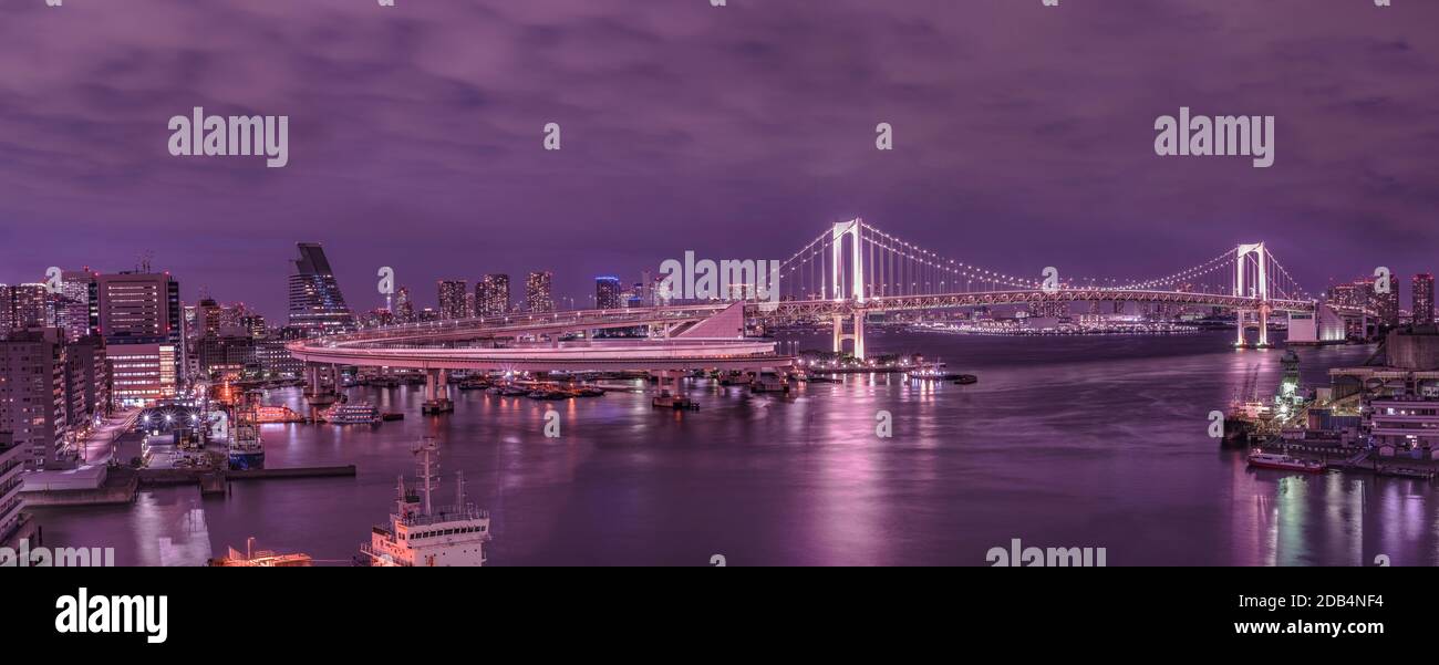 Panorama of the circular highway leading to the illuminated Rainbow Bridge with Cargo and cruise ships moored or sailing in Odaiba Bay of Tokyo. Stock Photo