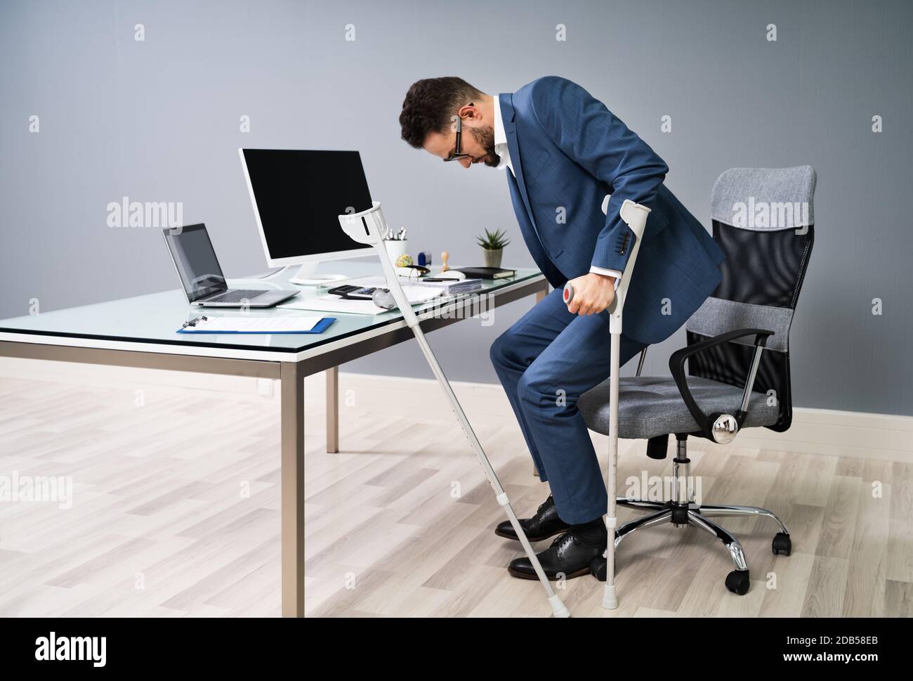 Man With Broken Leg Using Crutches To Get Up From Chair In Office Stock