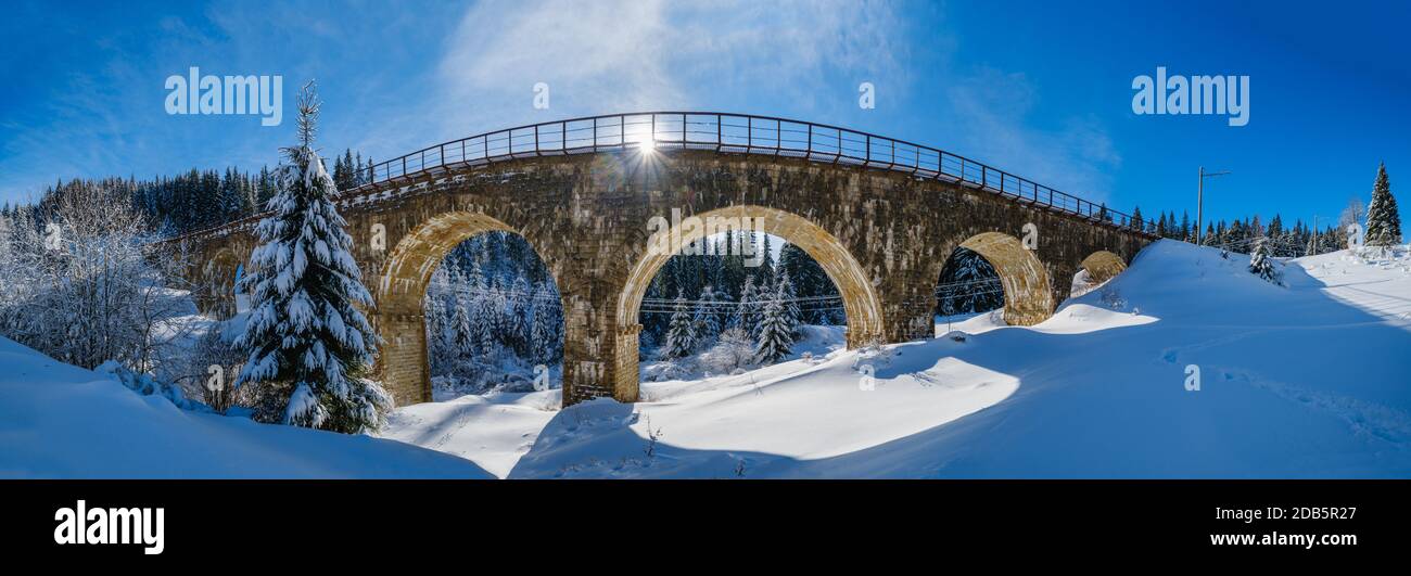 Stone viaduct (arch bridge) on railway through mountain snowy fir forest. Snow drifts  on wayside and hoarfrost on trees and electric line wires. Stock Photo