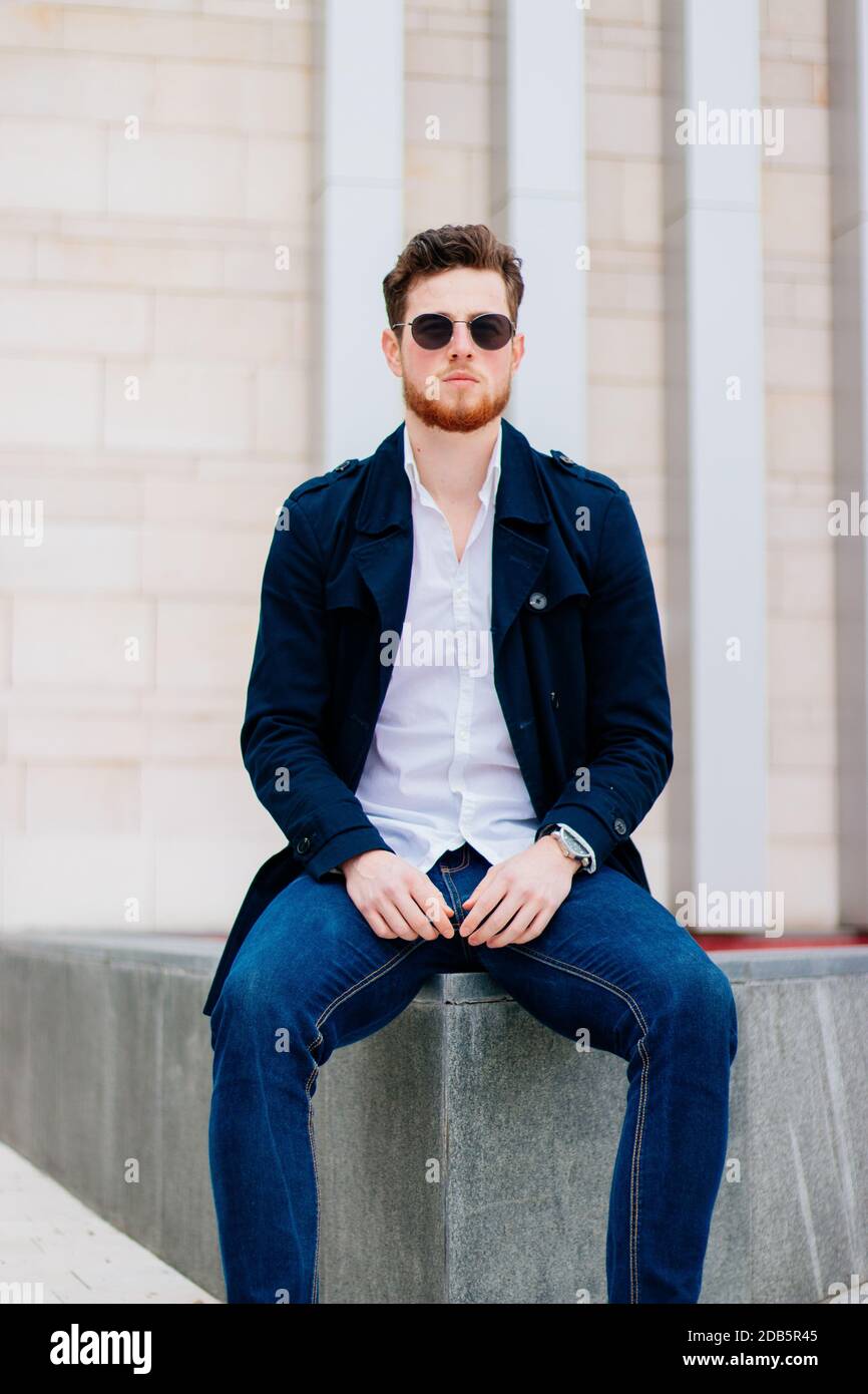 A Caucasian male wearing jeans pants, a white shirt, black coat and  sunglasses sitting on a wall Stock Photo - Alamy