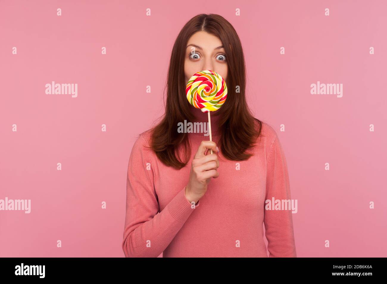 Surprised brunette woman in pink sweater licking lollipop and looking at camera with amazed expression. Indoor studio shot isolated on pink background Stock Photo
