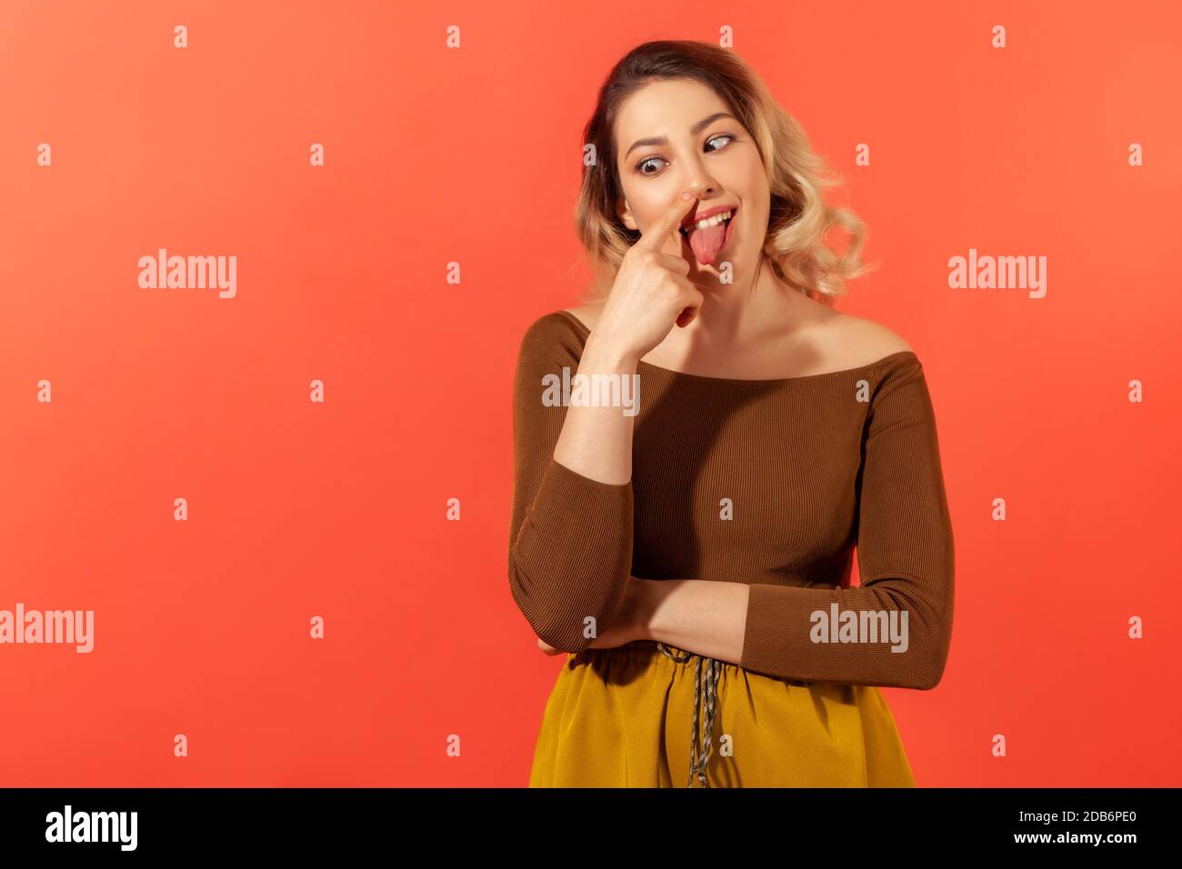 Portrait of funny woman in casual clothes touching her nose with forefinger, crossed eyes and tongue out. Funny and foolish concept. Indoor studio sho Stock Photo