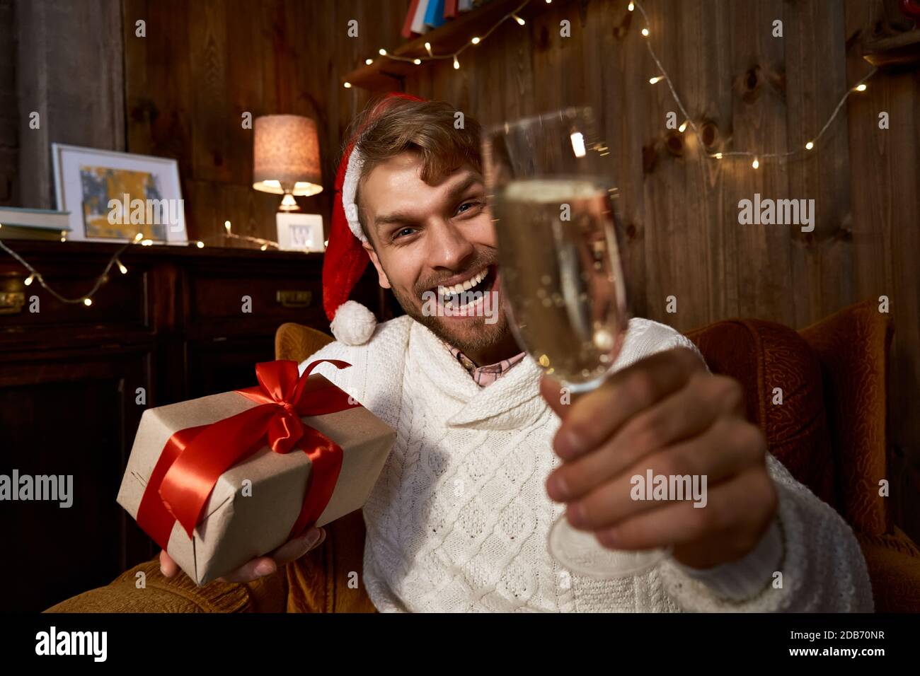 Happy guy wearing santa hat holding gift and champagne glass looking at web cam. Stock Photo