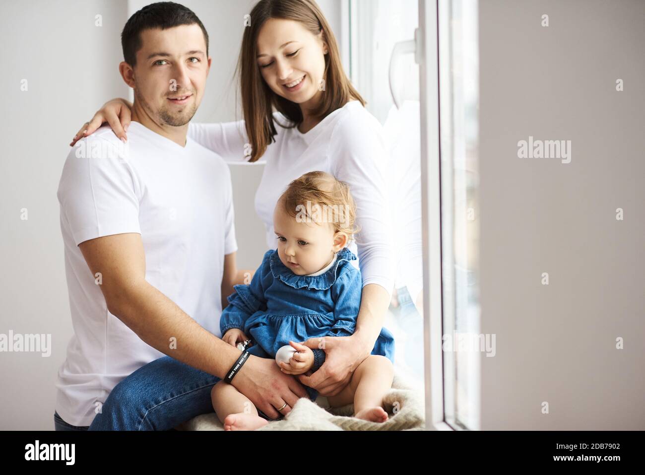 Portrait of happy young family with little daughter posing by the window in bright room. Stock Photo