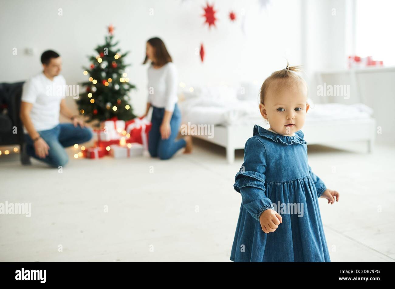 Portrait of a little baby girl dressed in a blue denim dress standing in a bright room. Stock Photo