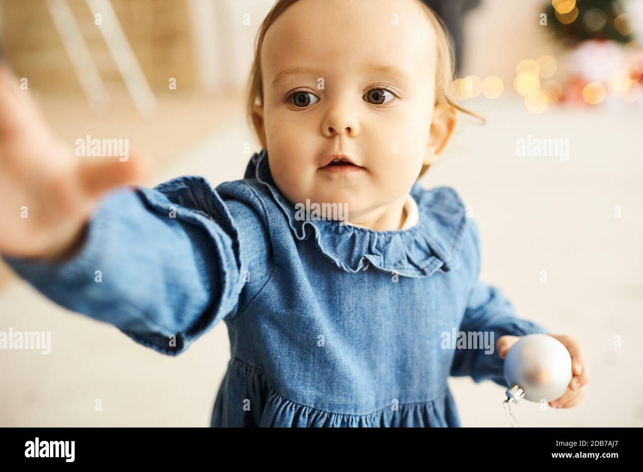 Portrait of a little baby girl dressed in a blue denim dress standing in a bright room. Stock Photo
