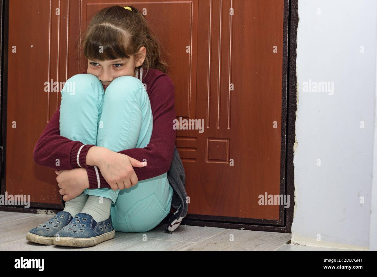 Upset girl sitting at the front door hugging her knees Stock Photo