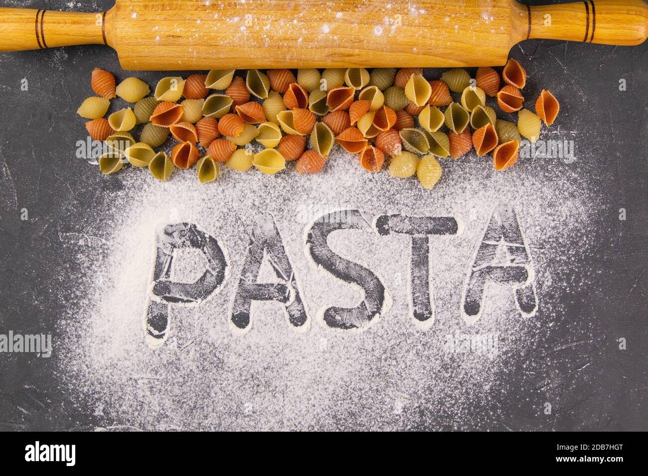 The words PASTA drawn a finger on flour with a kind of pasta, conchiglie and rocking for the dough. Italian pasta, flat lay close-up. Stock Photo