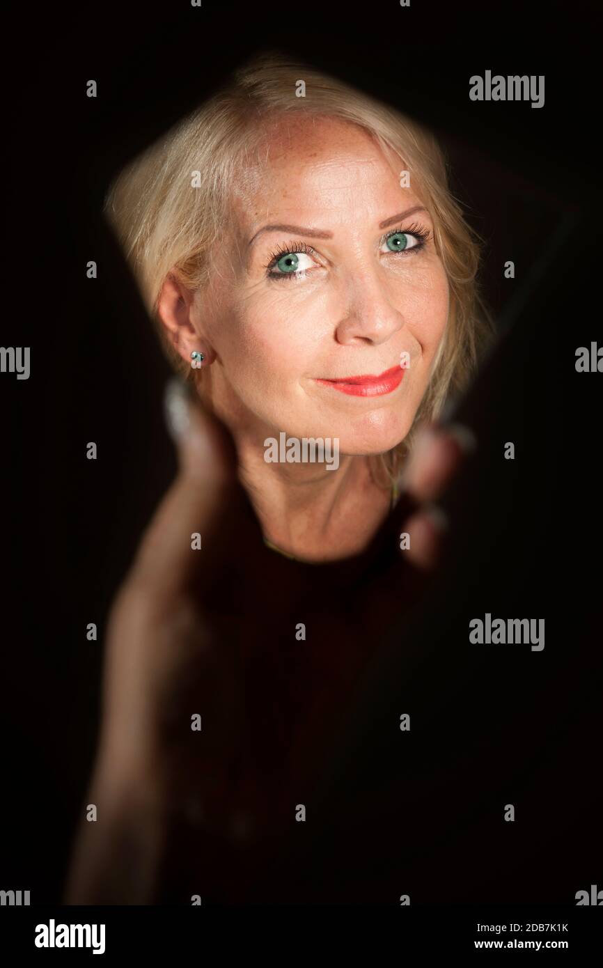 Mirror image of the face of a middle-aged blonde woman in a hand held mirror shard against a black background. Stock Photo