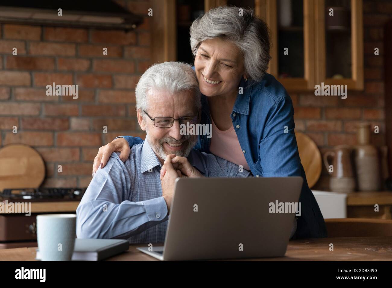 Smiling mature couple hugging, using laptop in kitchen together Stock Photo