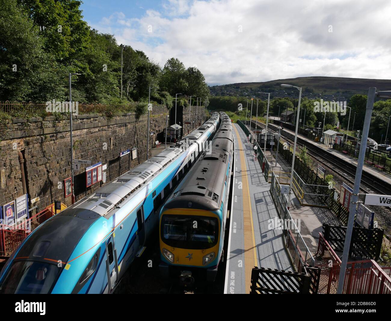 Two blue yellow and silver railway trains passing at railway station high stone wall and trees to left station platform to right hills in background Stock Photo