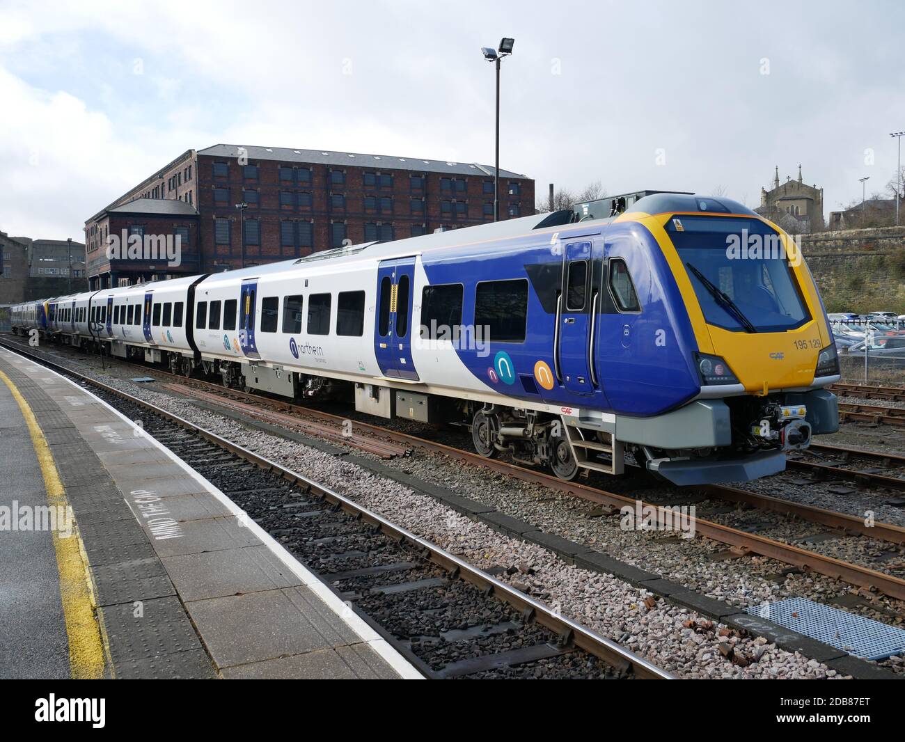 Yellow blue and silver multi carriage railway train parked at railway station red brick building in background Stock Photo