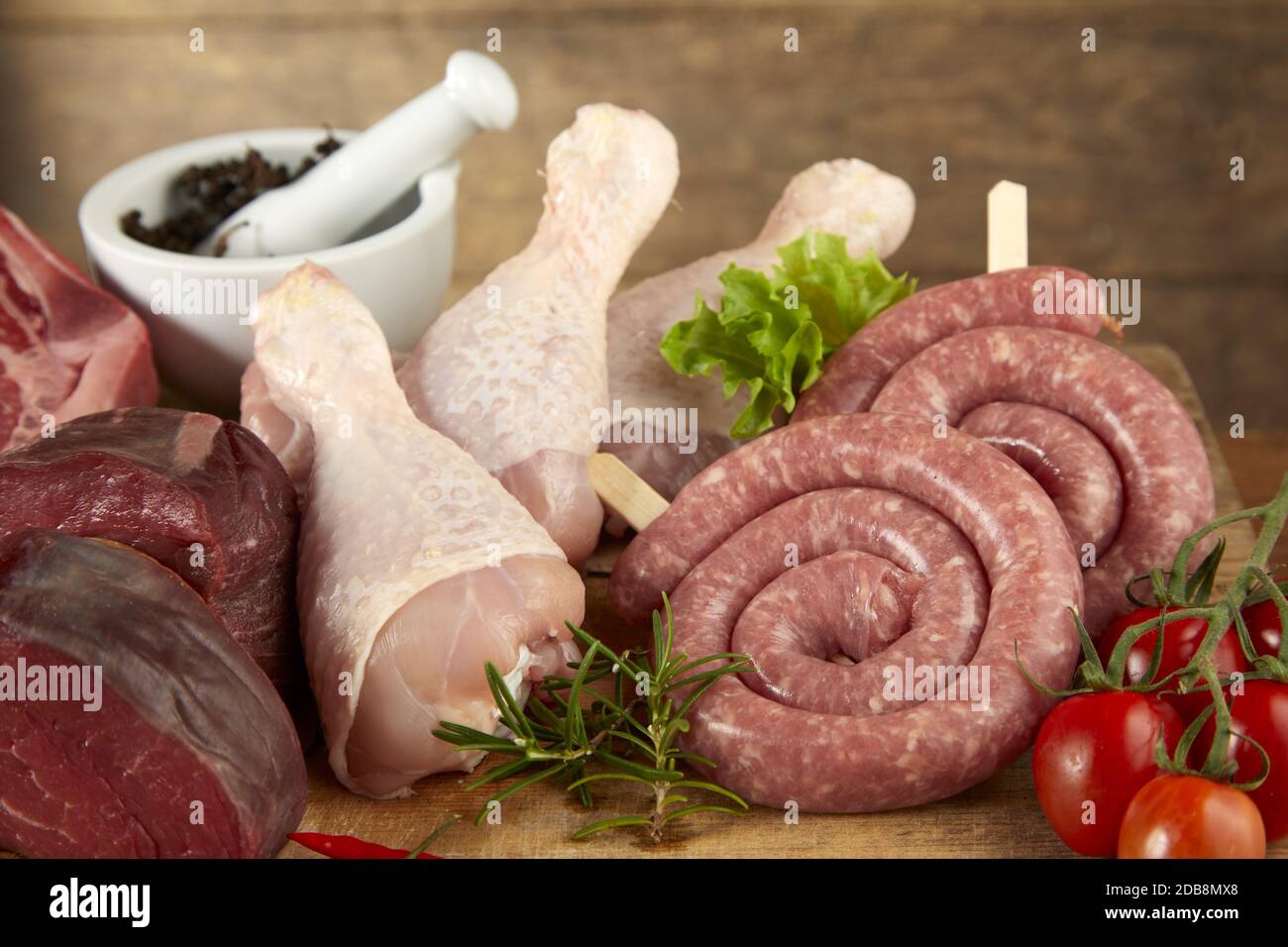 Assorted raw fresh meat for grilling with chicken legs, beef steak and sausage coils on a wooden background with herbs and spices in close up Stock Photo