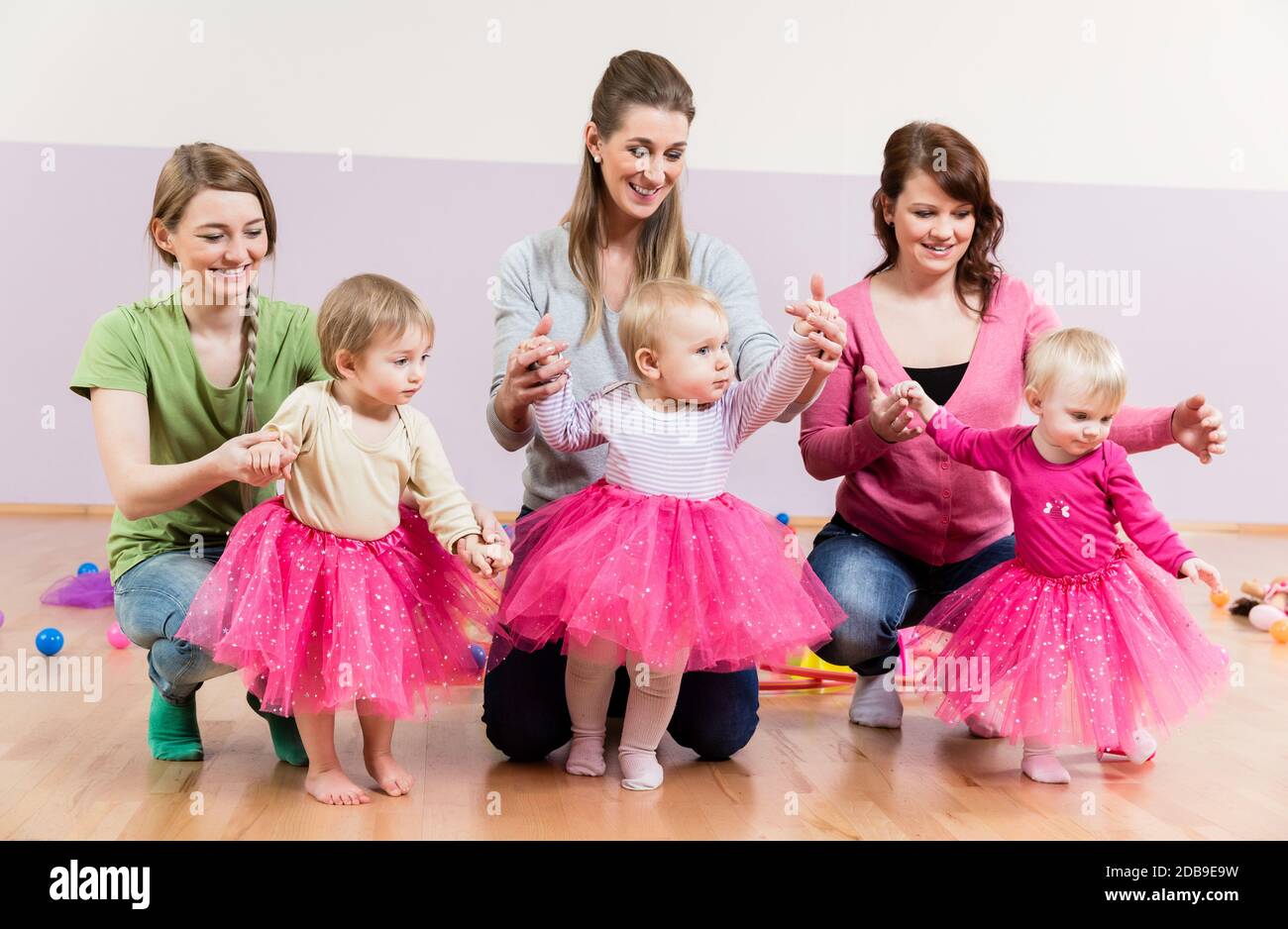 Three baby girls in pink skirts learning to walk with help of their mothers Stock Photo