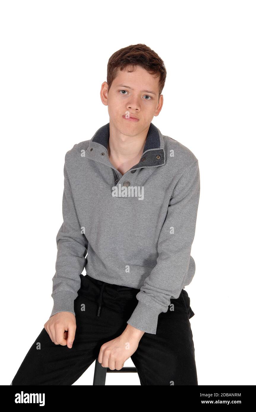 A happy young teenage boy sitting on a chair in the studio in a gray sweater, looking into the camera, isolated for white background Stock Photo
