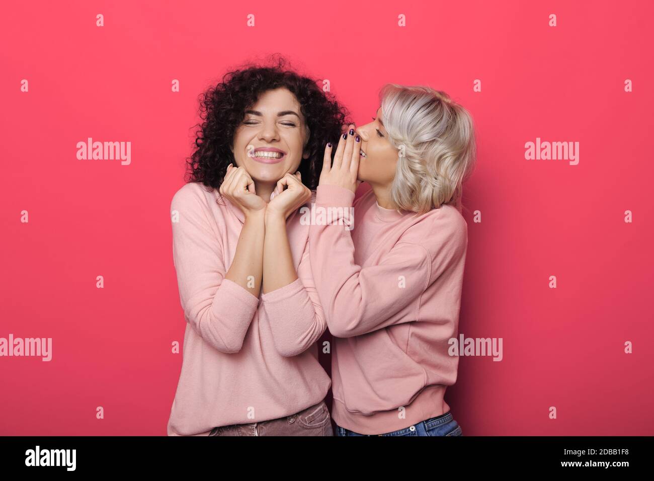 Adorable woman with blonde hair is whispering something to her brunette curly haired sister on red studio wall Stock Photo