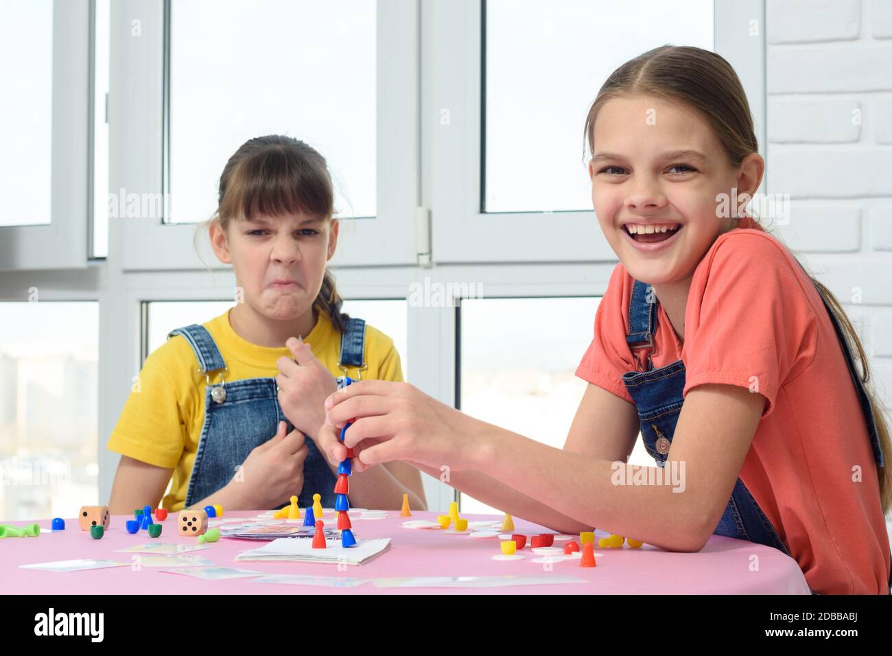 One girl laughs cheerfully, the other makes faces, children play a board game Stock Photo