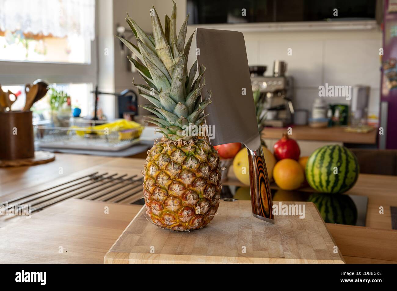 A cutting board with a whole pineapple fruit and a cleaver standing on a worktop in the kitchen in the background are more fruits and kitchen utensils Stock Photo