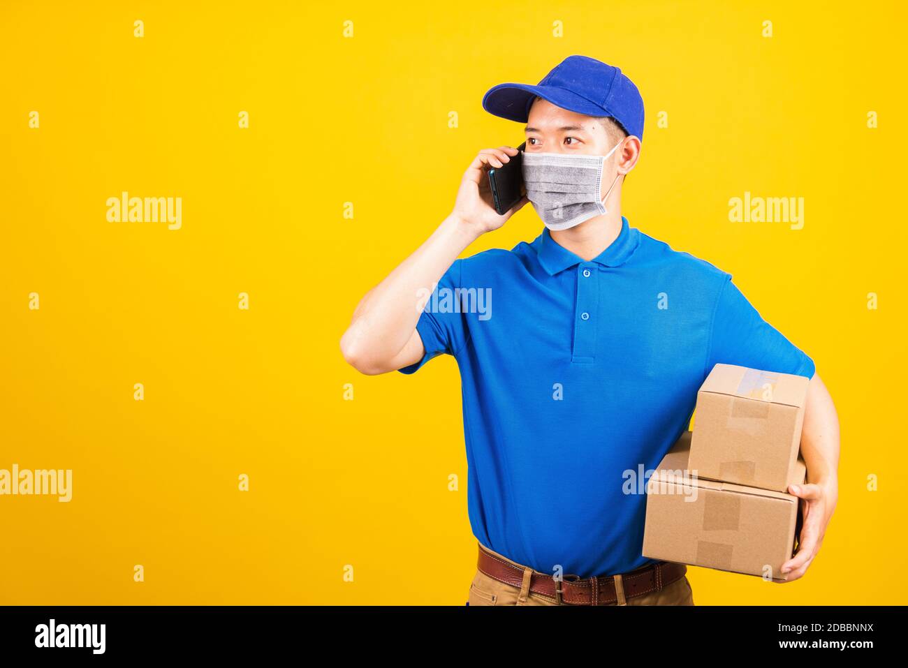 Asian young delivery worker man t-shirt and cap uniform wearing face mask protective use smart mobile phone call to customer under coronavirus or COVI Stock Photo