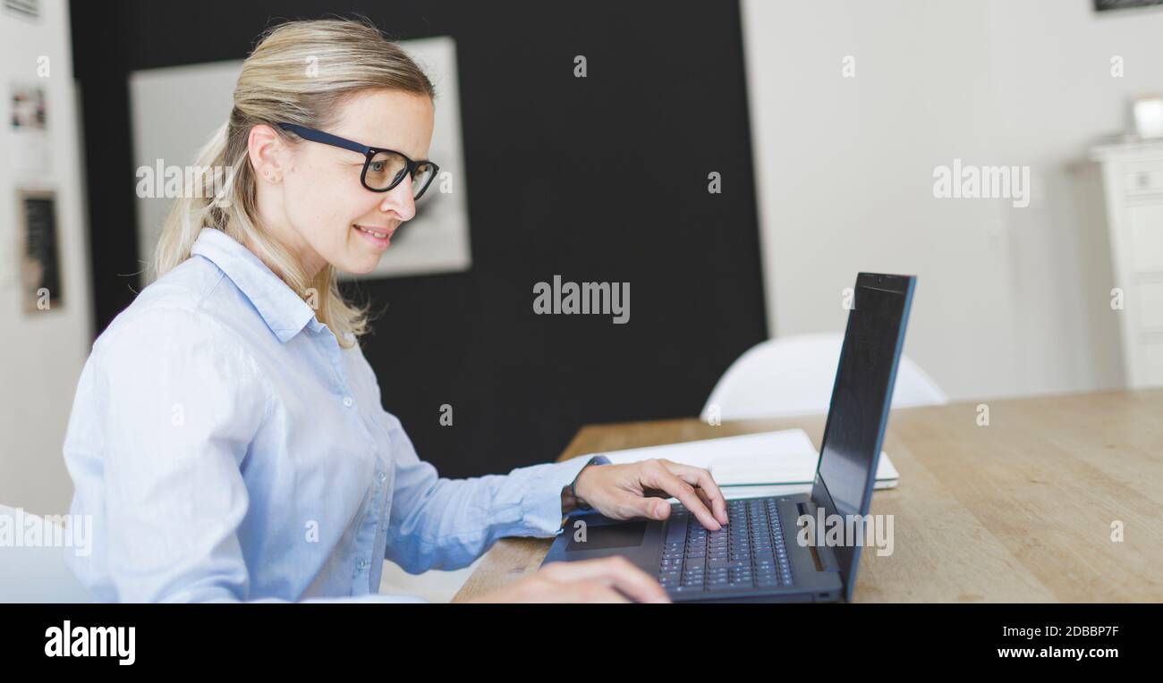 pretty young woman with blue shirt and glasses doing home office sitting at the kitchen table in front of her laptop Stock Photo