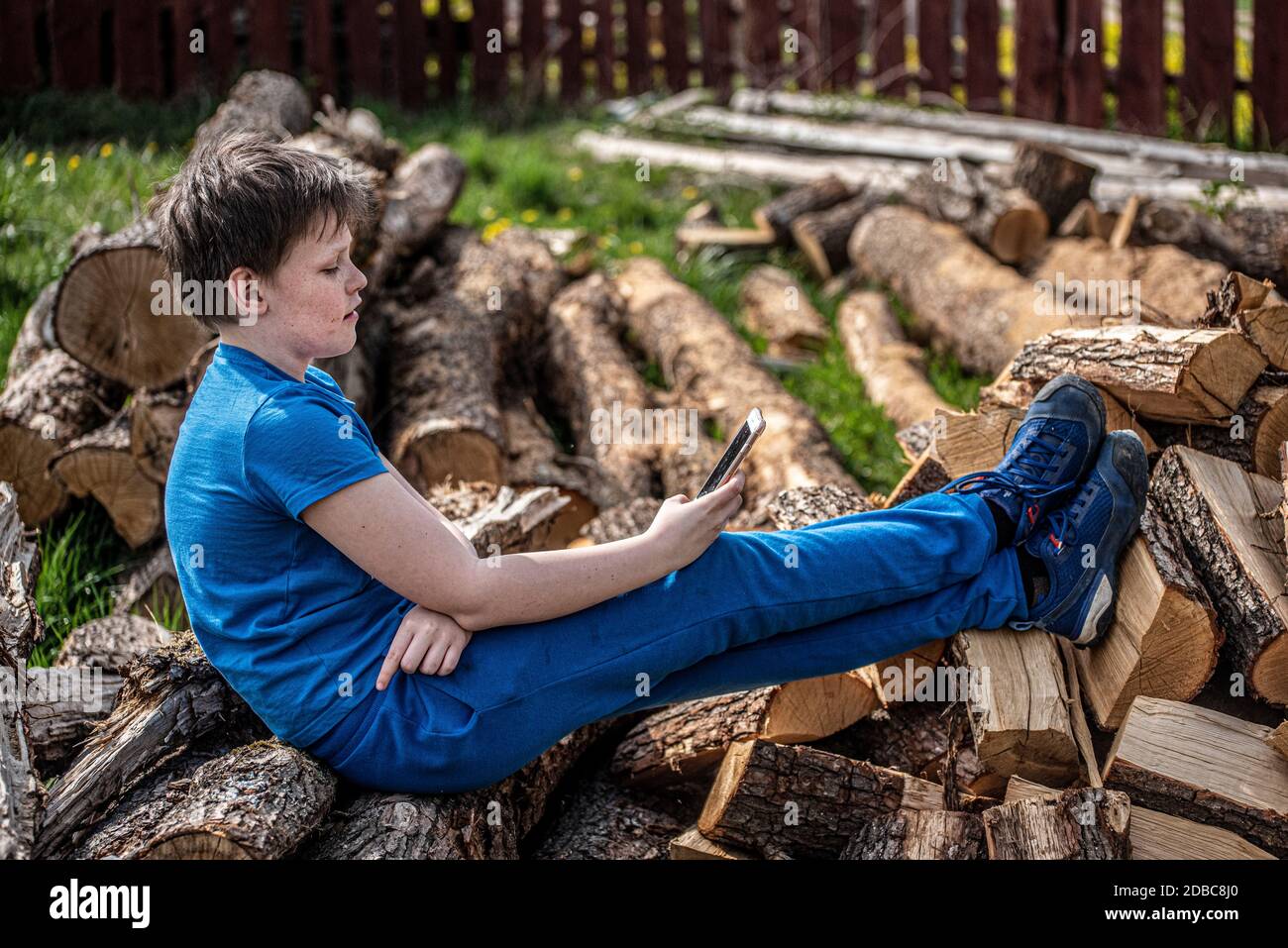 Boy sitting outdoors on a pile of firewood and looking at his mobile phone Stock Photo