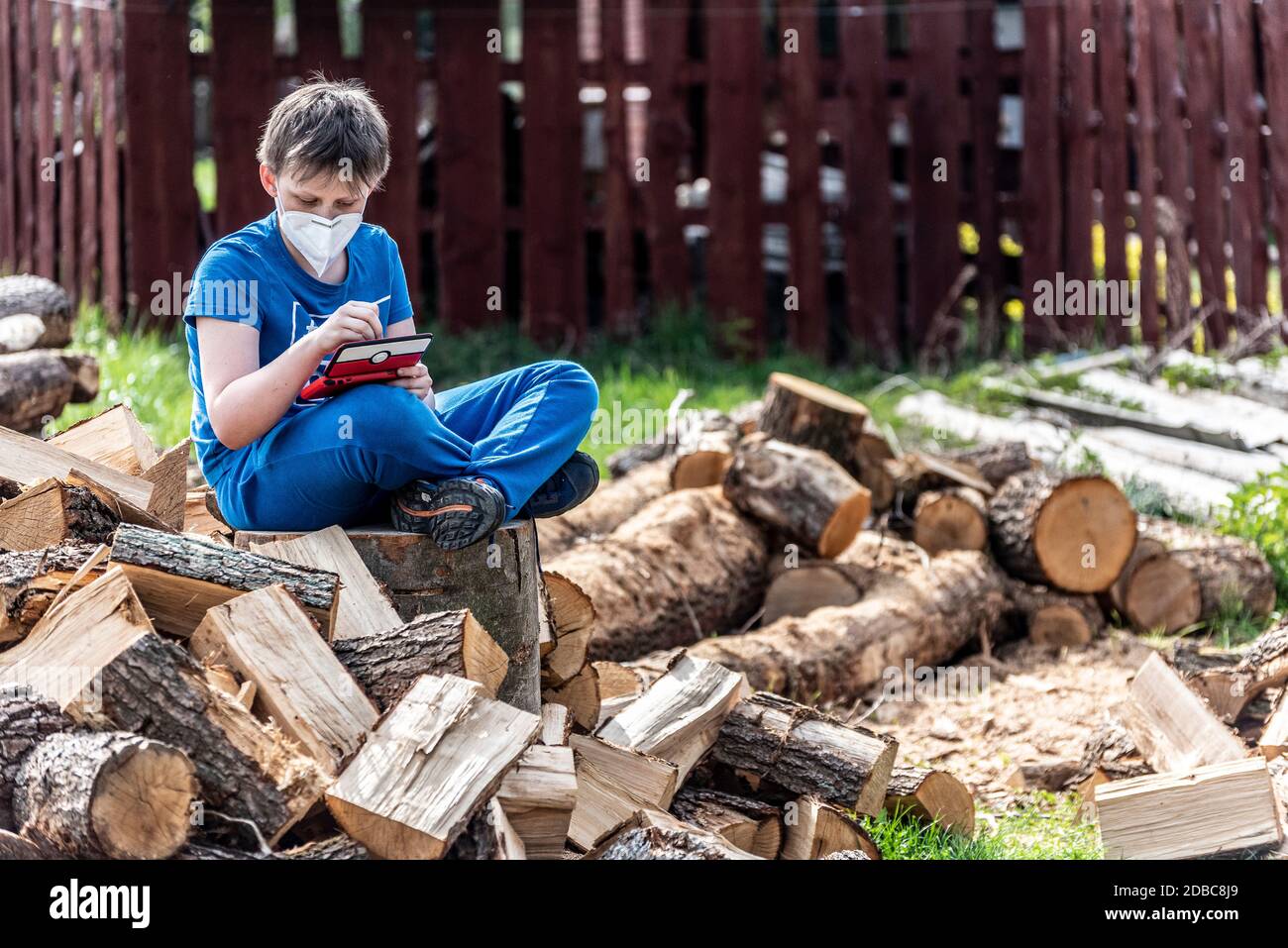 Boy with a protective face mask sitting on a pile of chopped firewood plays with a Nintendo switch console Stock Photo