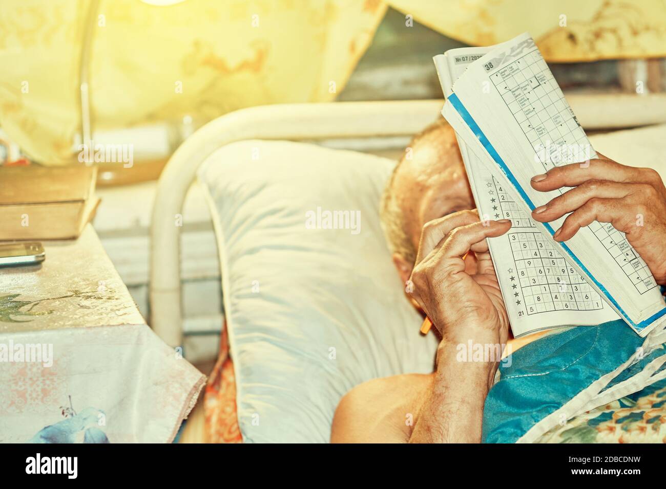 Senior man doing crossword puzzle lying in bed by lamp light Stock
