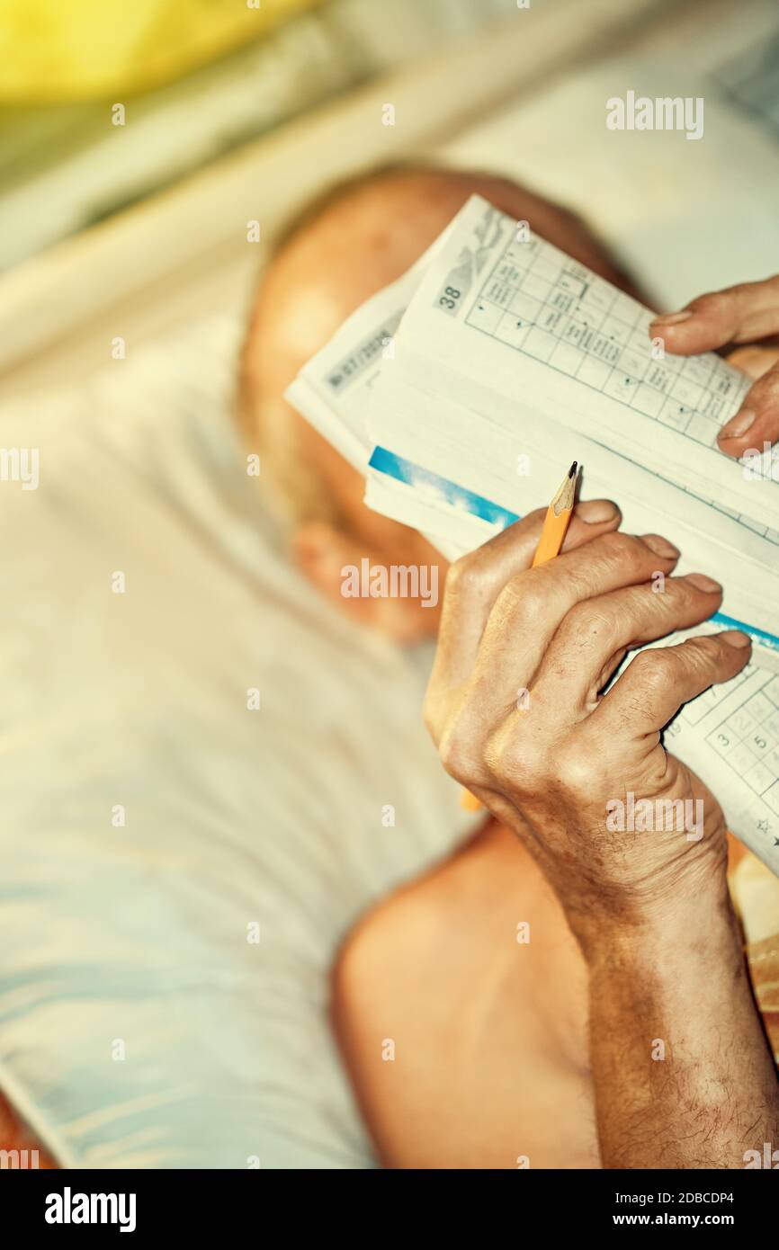 Senior man doing crossword puzzle lying in bed by lamp light Stock