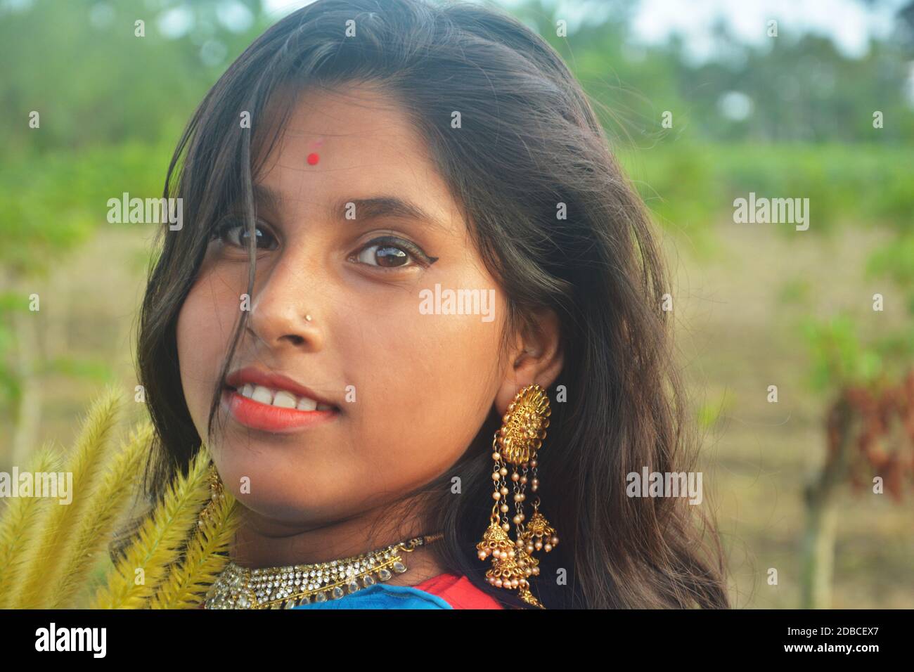 Close up of a Indian Bengali smiling teenage girl with long dark hairs golden colored wearing earrings and necklace with a red bindi on her forehead, Stock Photo