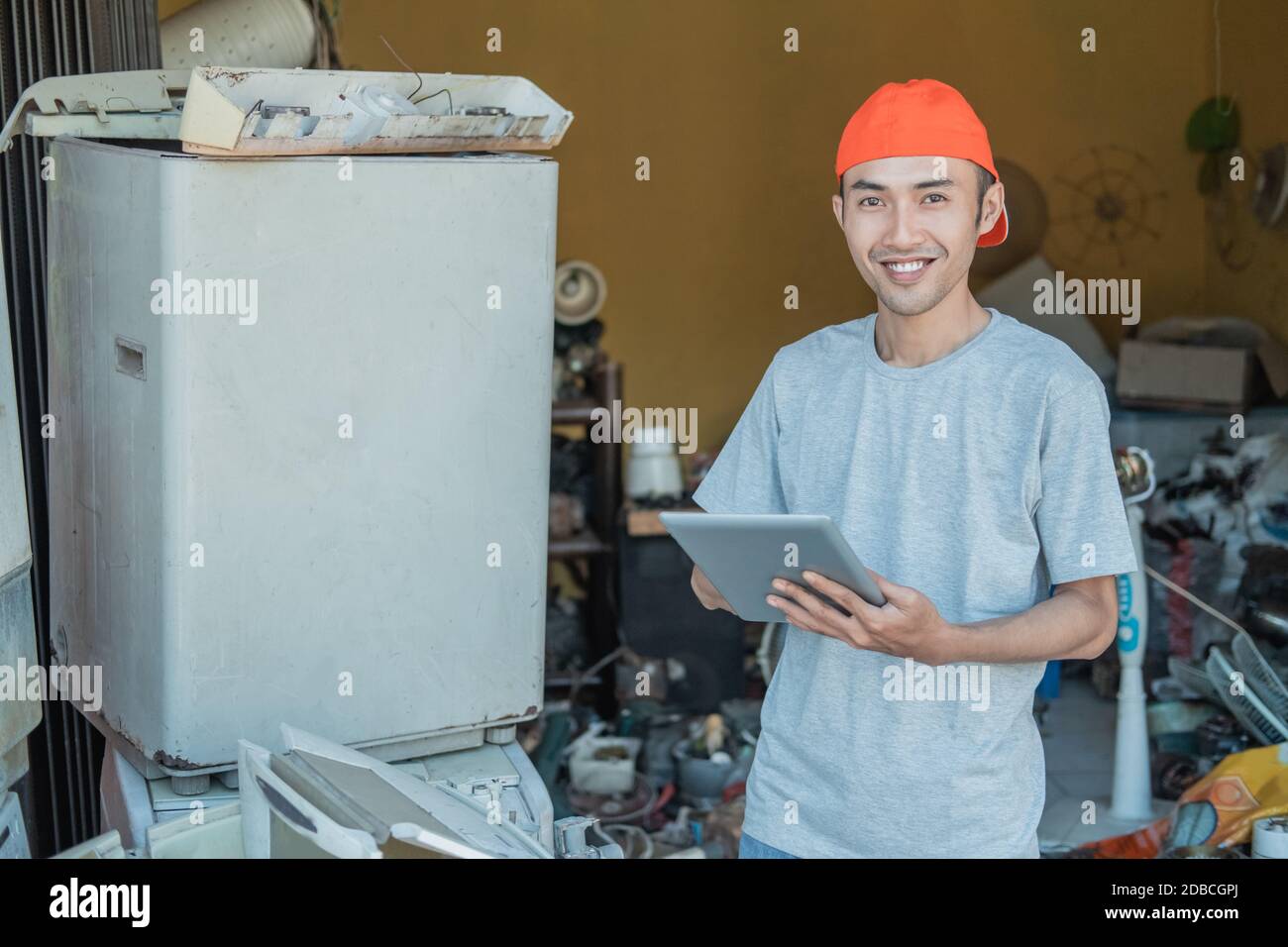 asian electronics repairman smiles at the camera while using his tablet pc around broken electronics in service shop Stock Photo